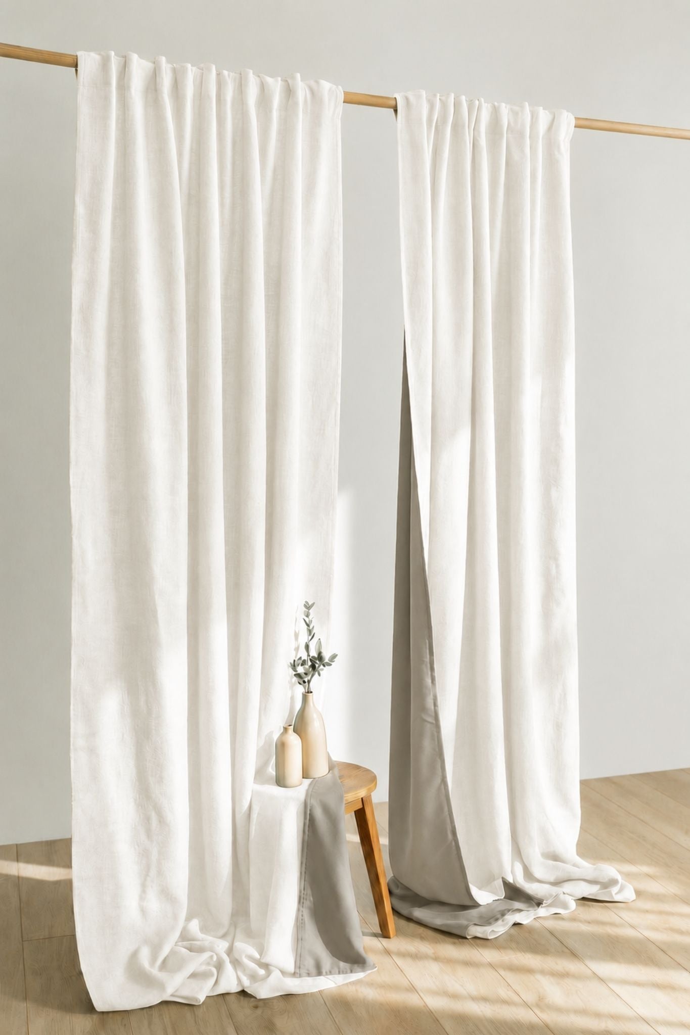 Two long off white curtains hanging from a wooden rod in a bright room with light wood flooring. A small stool with two beige vases stands near the floor, and sunlight streams across the scene.