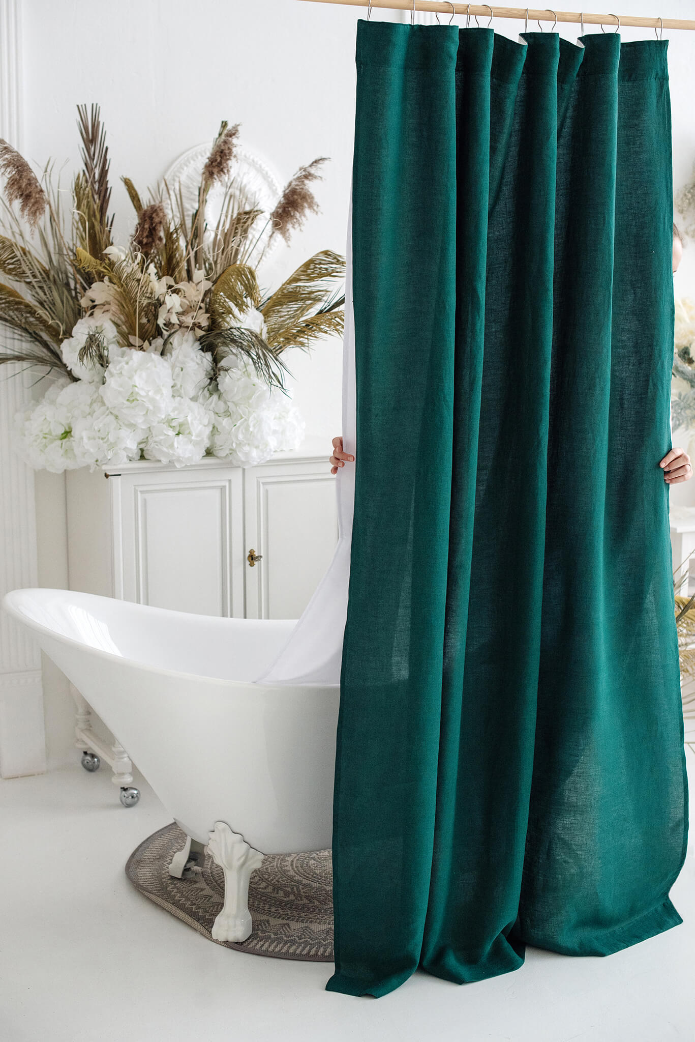 A dark teal green curtain panel hanging from a wooden rod in a bright bathroom setting next to a white clawfoot tub and dried floral decor.