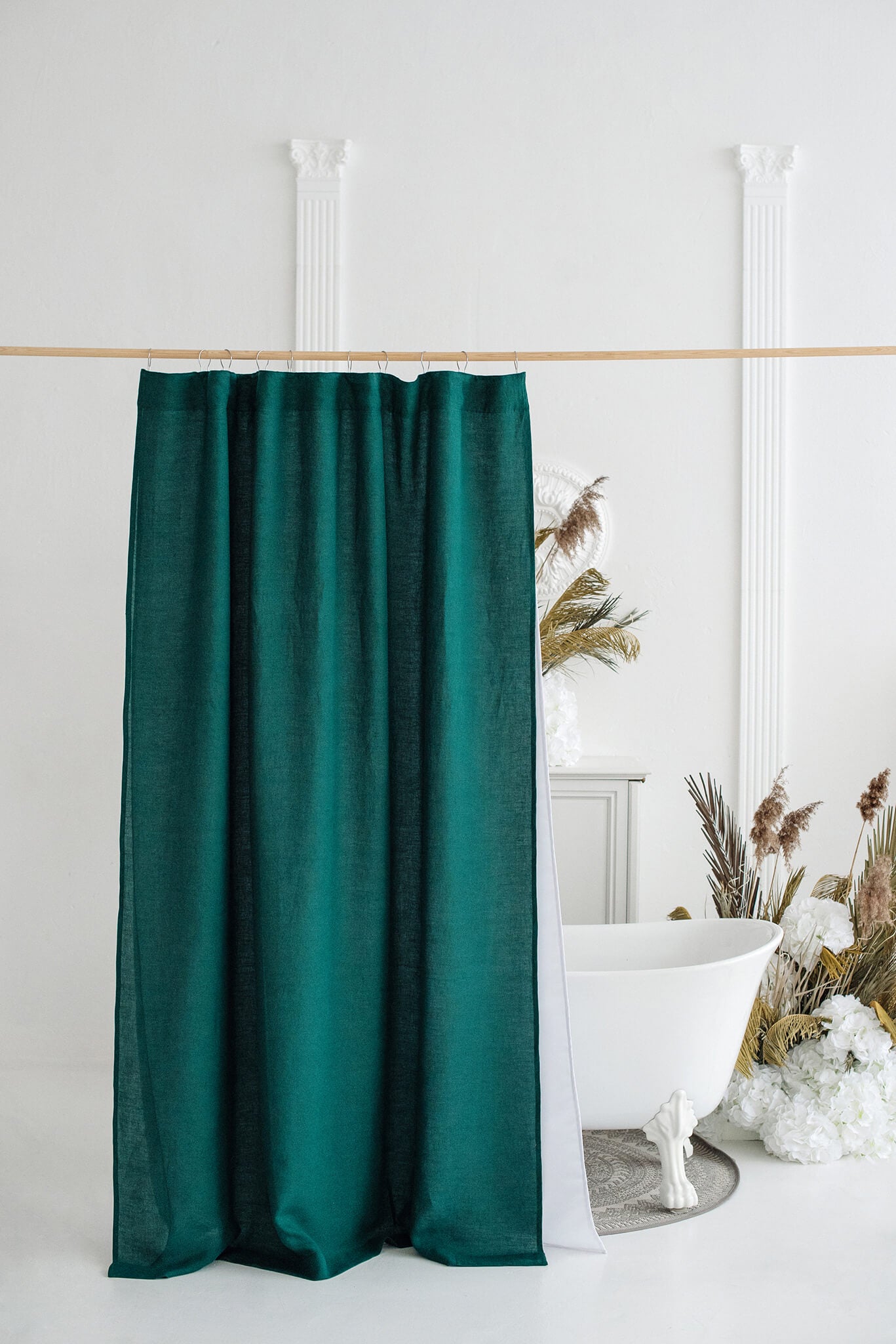 A dark teal green velvet shower curtain hanging from a light wood rod in a bright, white bathroom setting with a clawfoot tub and dried pampas grass decor.