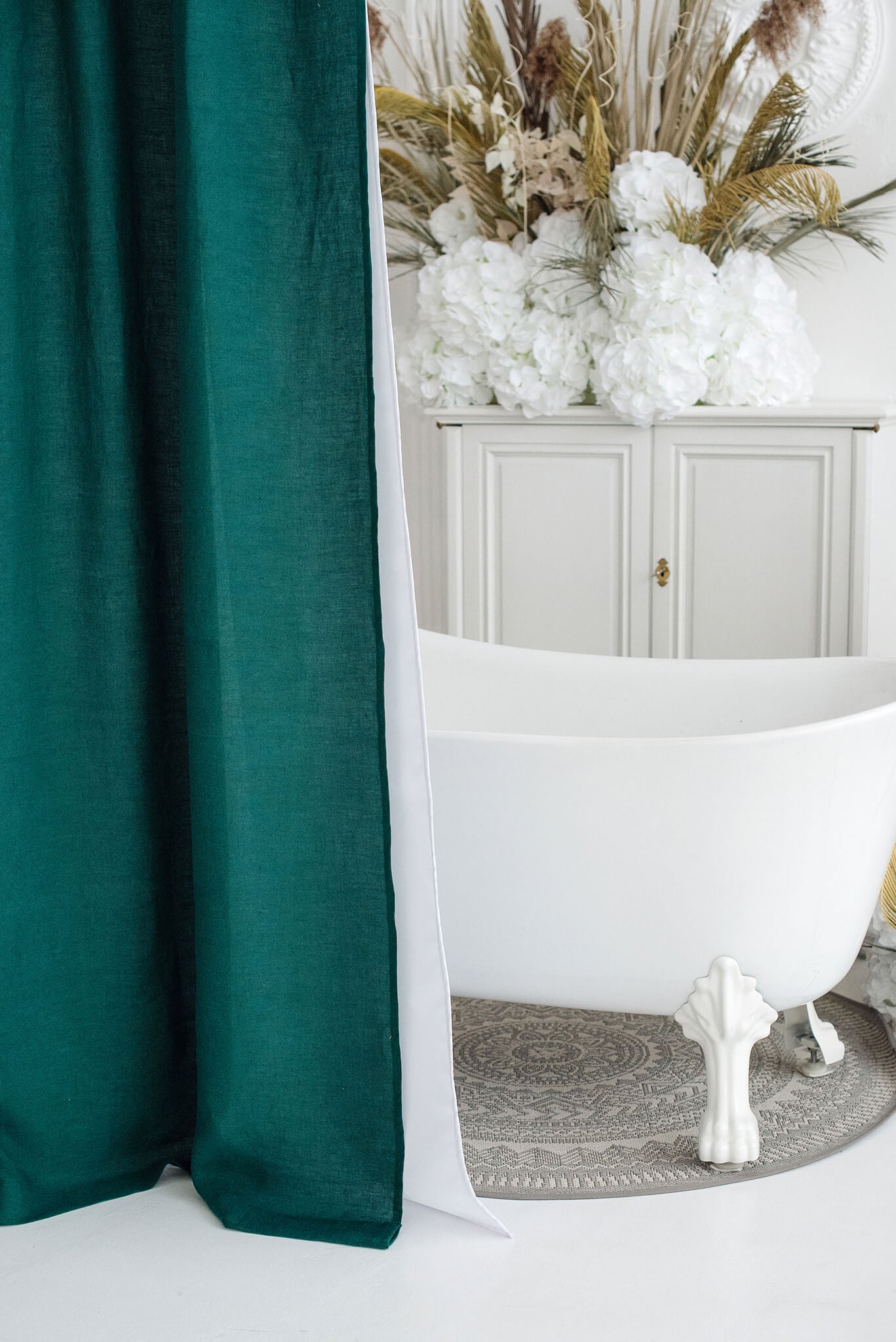 A dark teal green velvet shower curtain hanging next to a white freestanding clawfoot bathtub in a styled bathroom setting with a white cabinet and dried floral arrangements in the background.