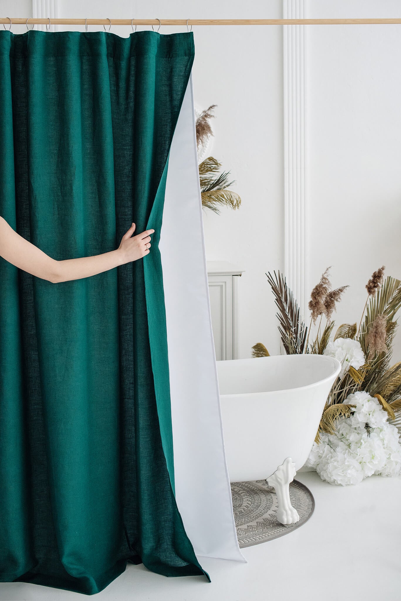 A person's arm is shown pulling a dark green velvet shower curtain to reveal a white shower screen inside a bright, modern bathroom with a clawfoot tub and dried pampas grass decor.