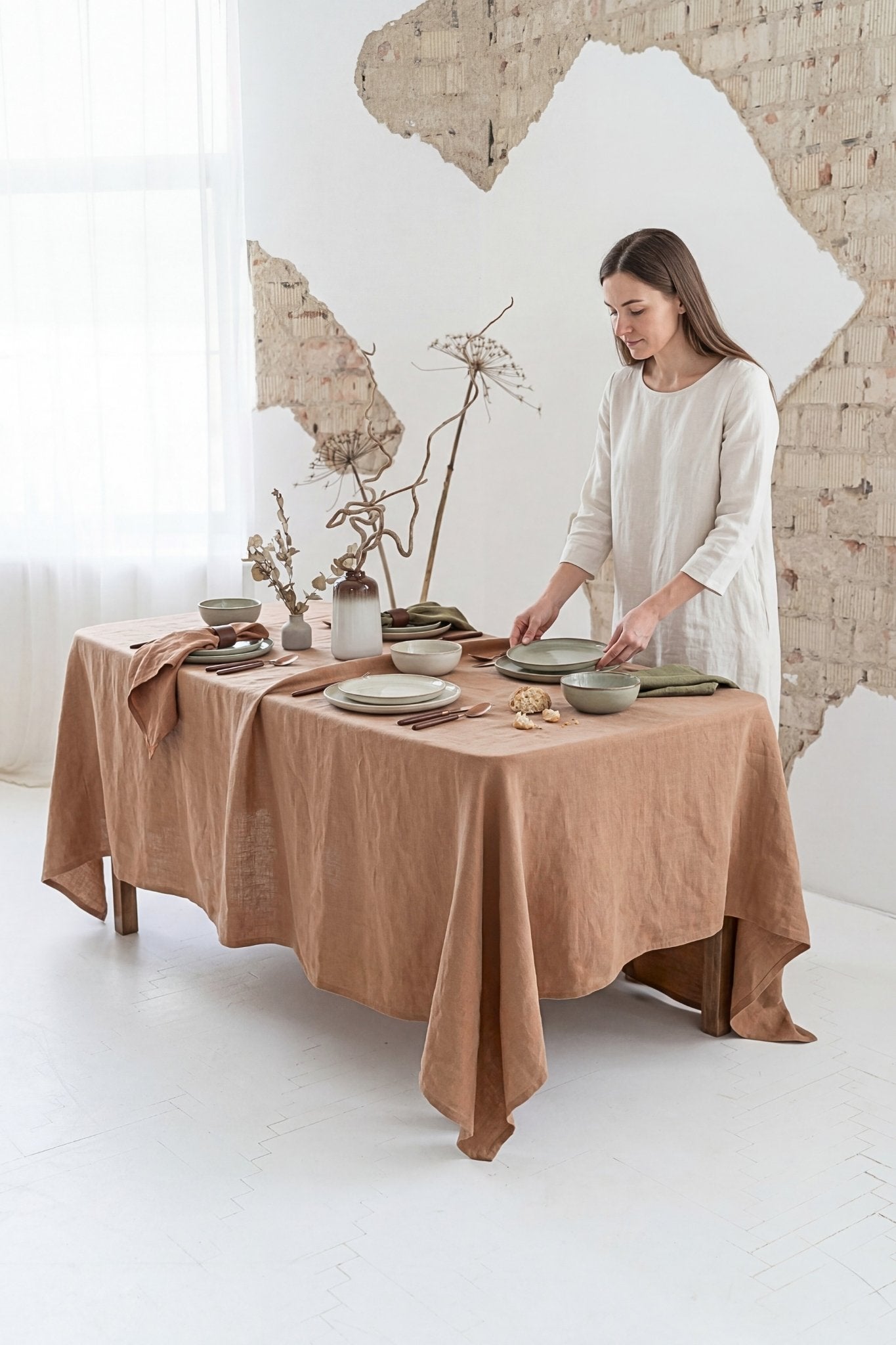 Woman setting a table with a brown linen tablecloth in a room with a brick wall.