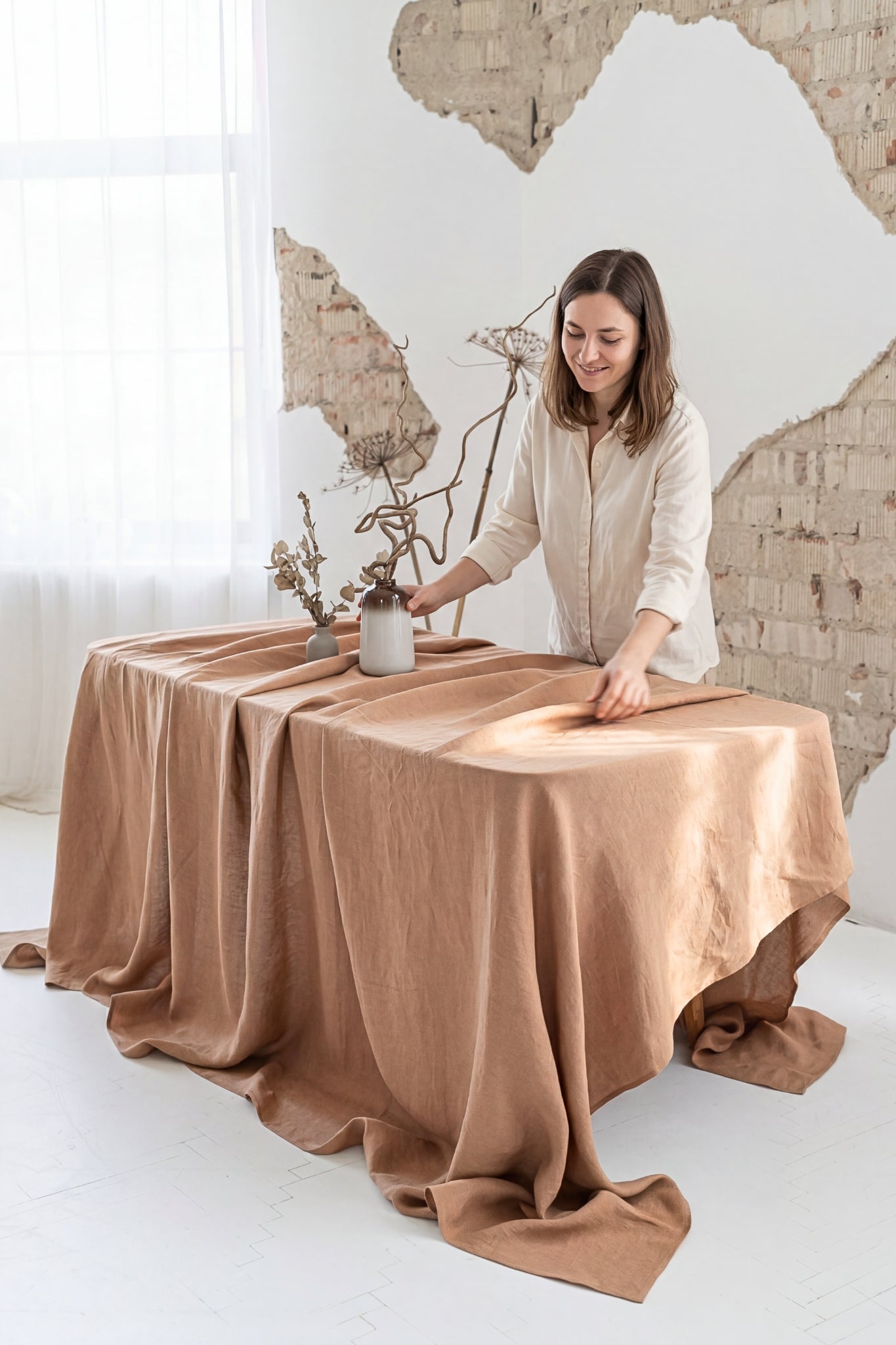 Woman arranging items on a table draped with a brown linen tablecloth against a white wall.
