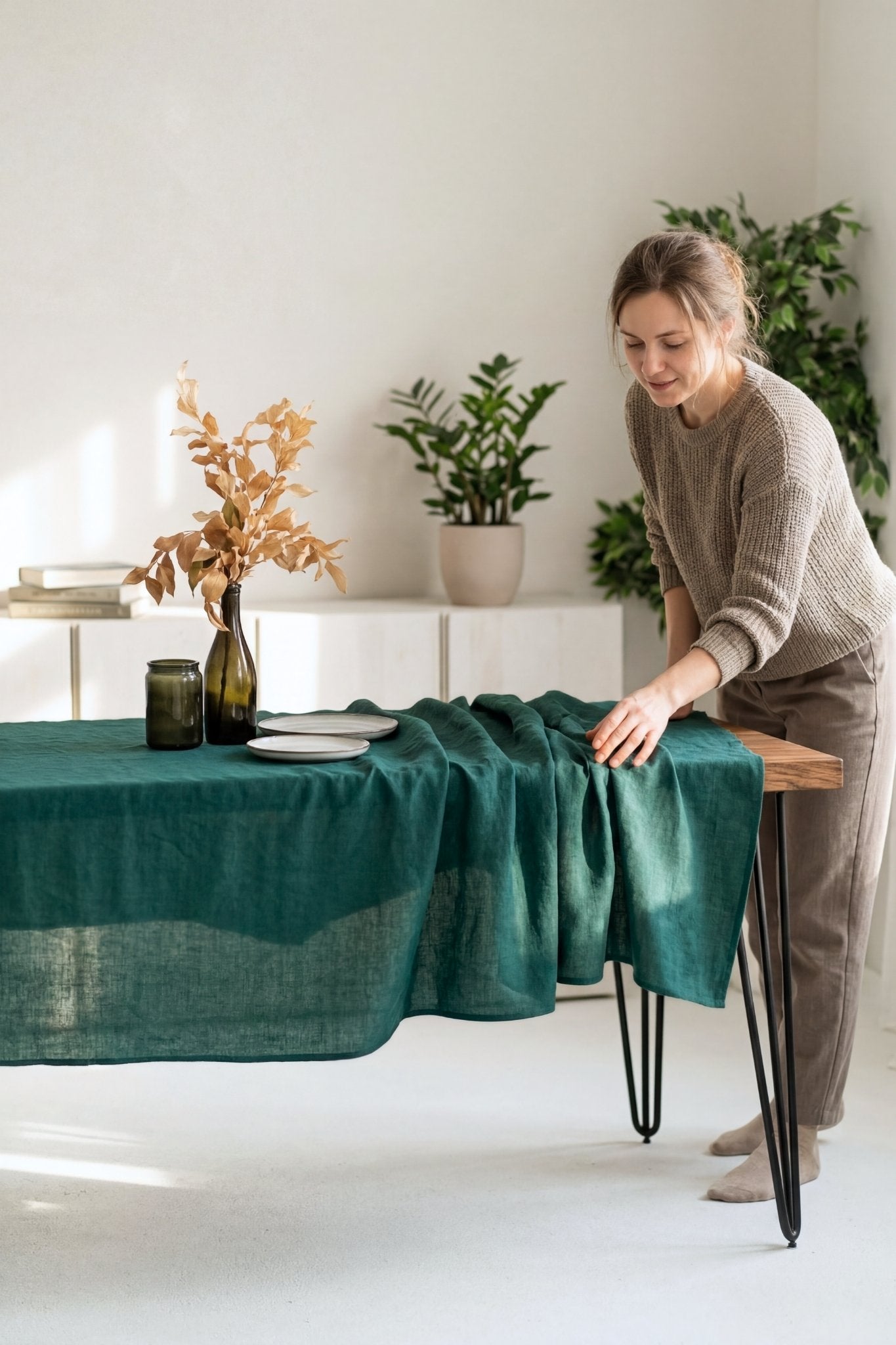 Woman arranging a green linen tablecloth on a dining table in a bright room with plants.