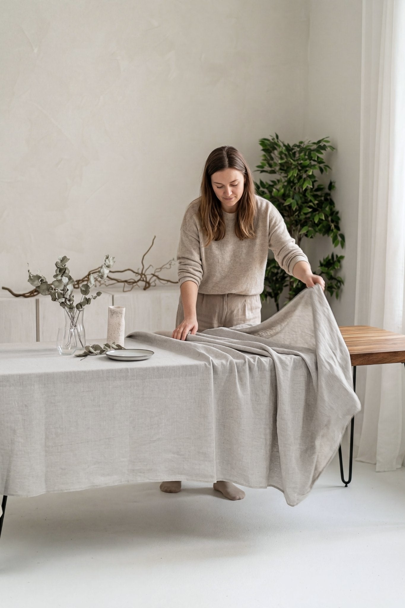 Woman arranging a table with a neutral linen tablecloth in a minimalistic room.