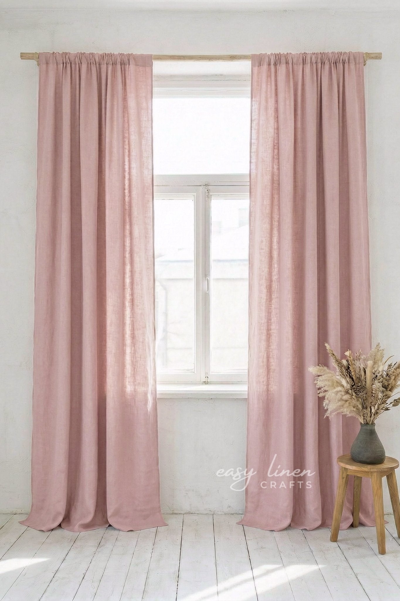 Pink curtains hanging in a window with a white wall and wooden stool with flowers in the background.