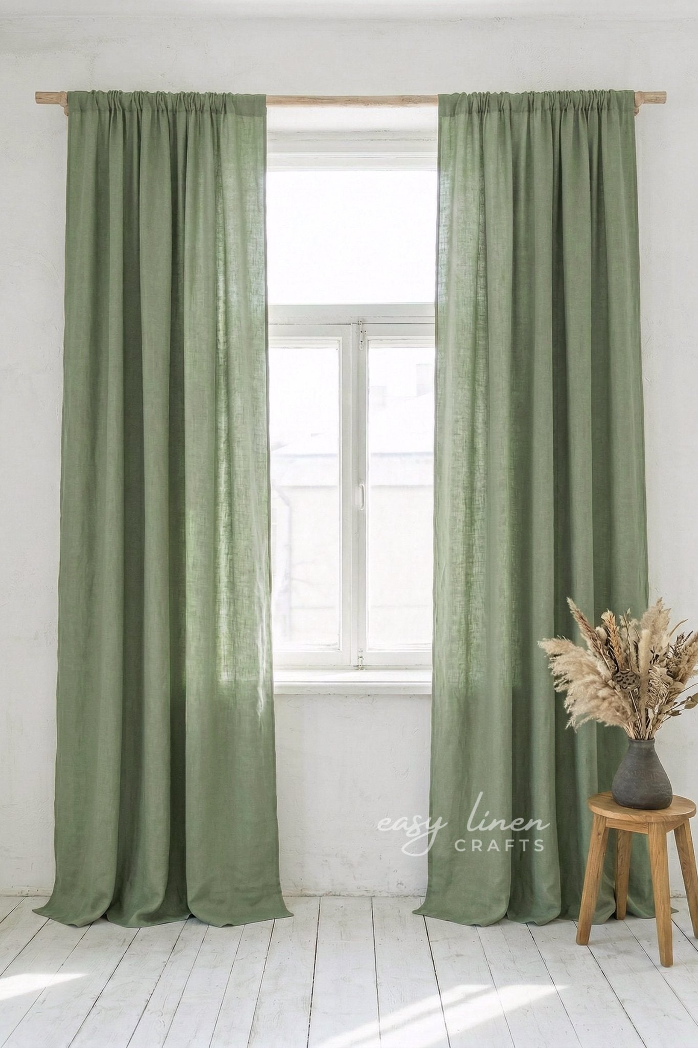 Green curtains hanging in a window with a white wall and wooden stool with dried plants in the background.