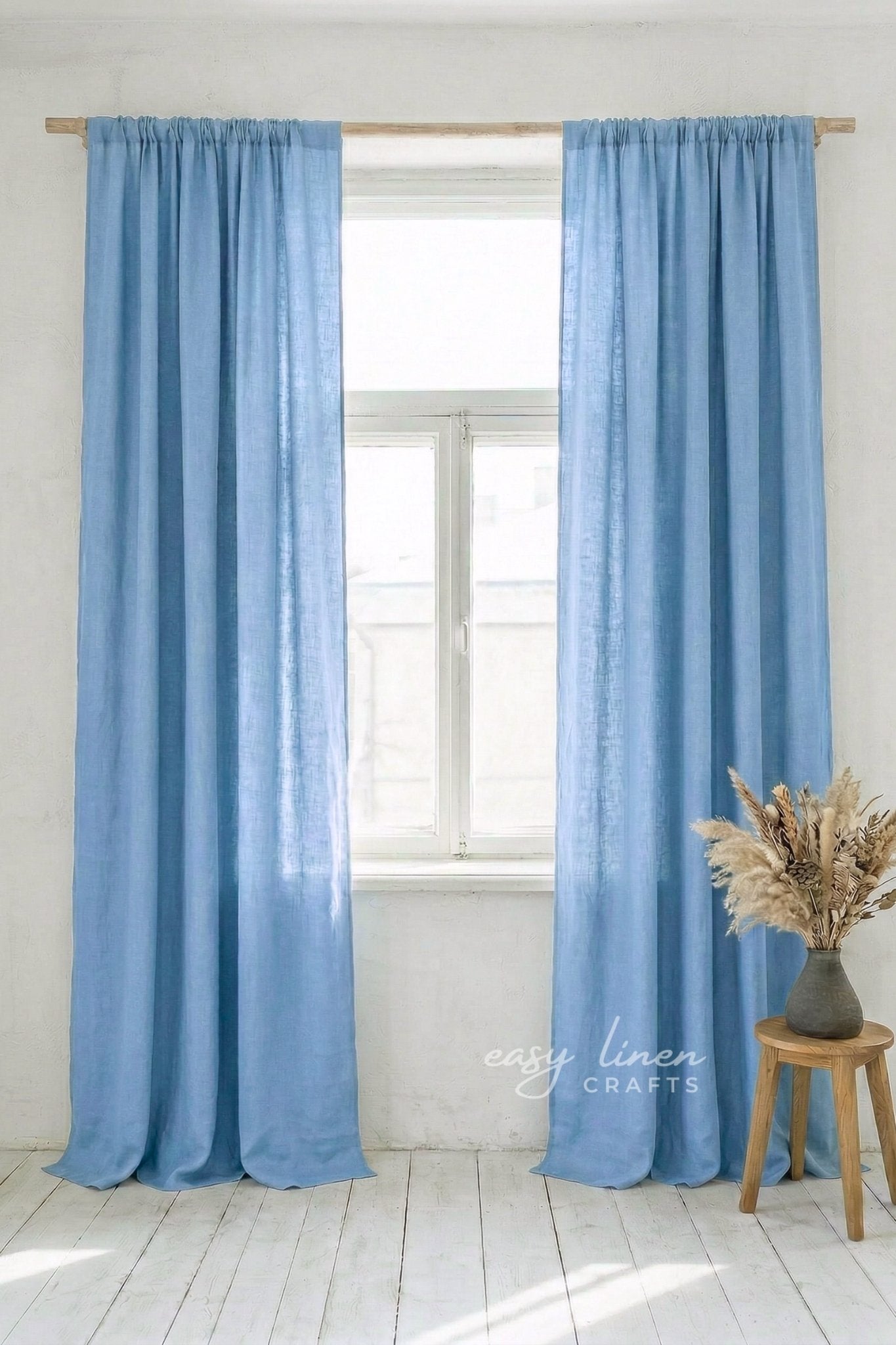 Light blue curtains hanging in a window with a white wall and wooden stool with dried plants in the background.