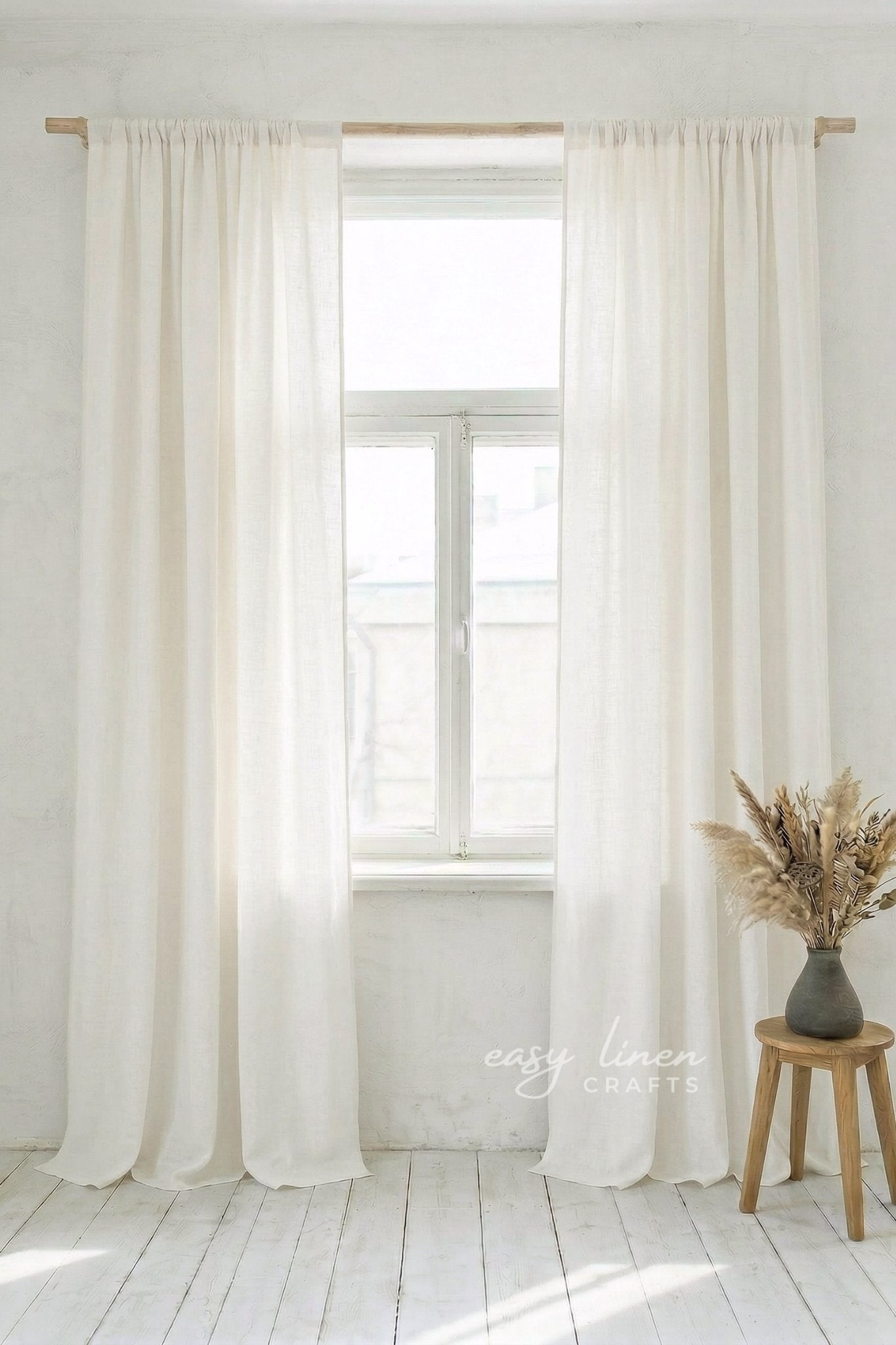 White curtains hanging in a window with a wooden stool and vase of dried flowers on a white floor.