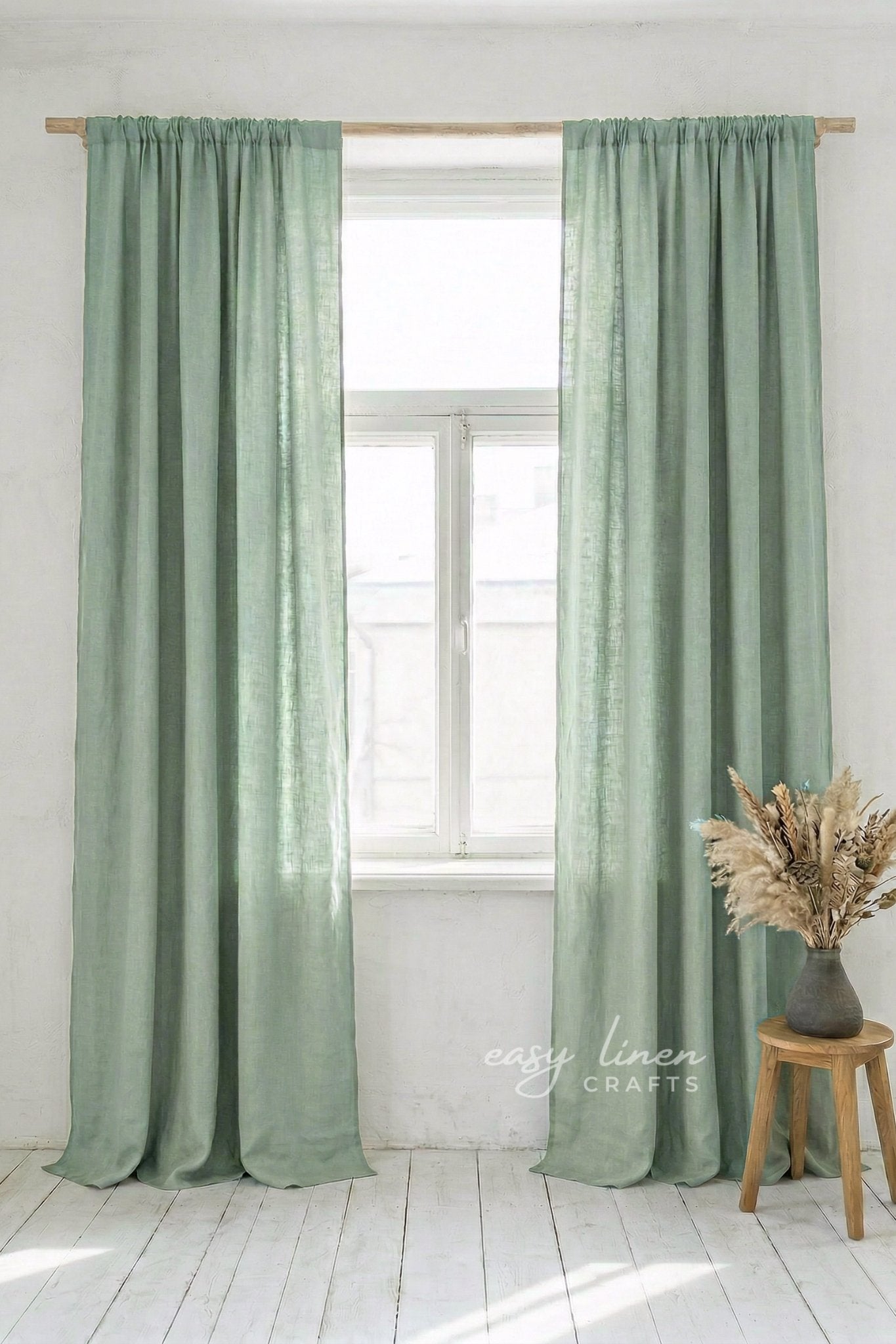 Green curtains hanging in a window with a white wall and wooden stool with dried plants in the background.