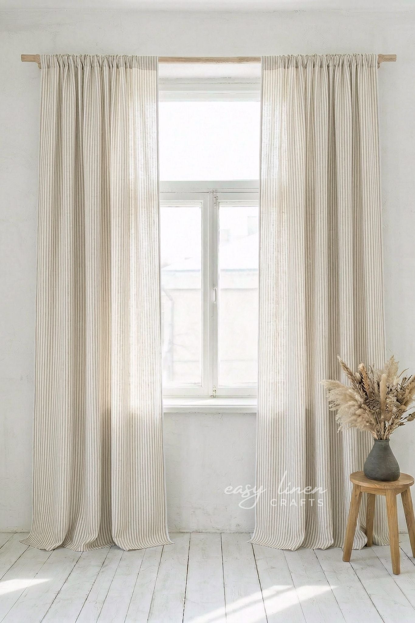 Striped curtains hanging in a window with a wooden stool and vase of dried flowers on a white floor.