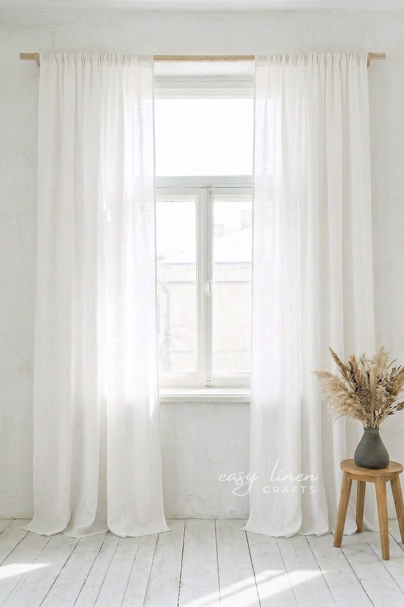 White curtains hanging in a bright room with a wooden stool and vase of dried flowers.