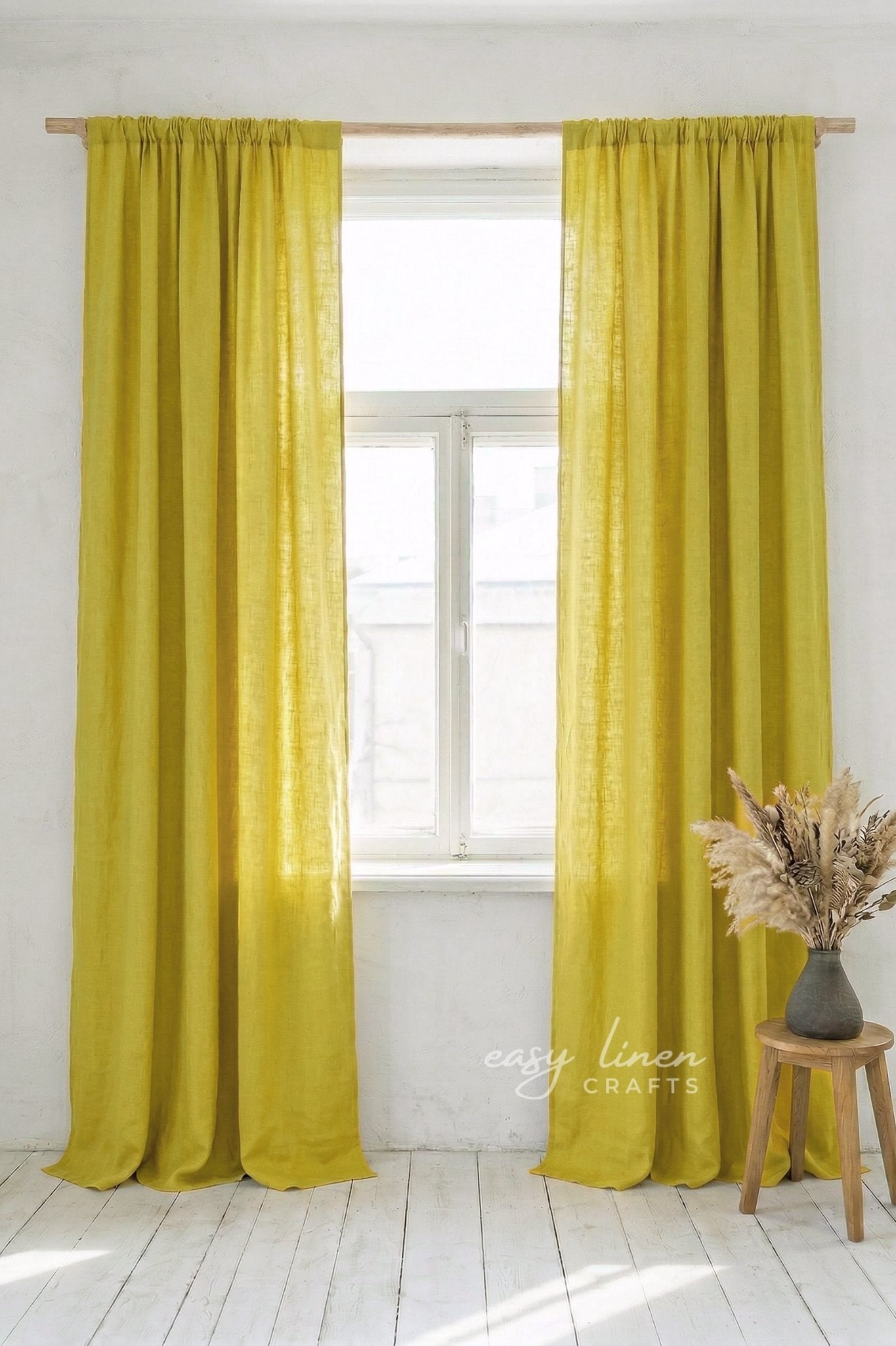 Yellow curtains hanging in a window with a white wall and wooden stool with dried plants in the foreground.