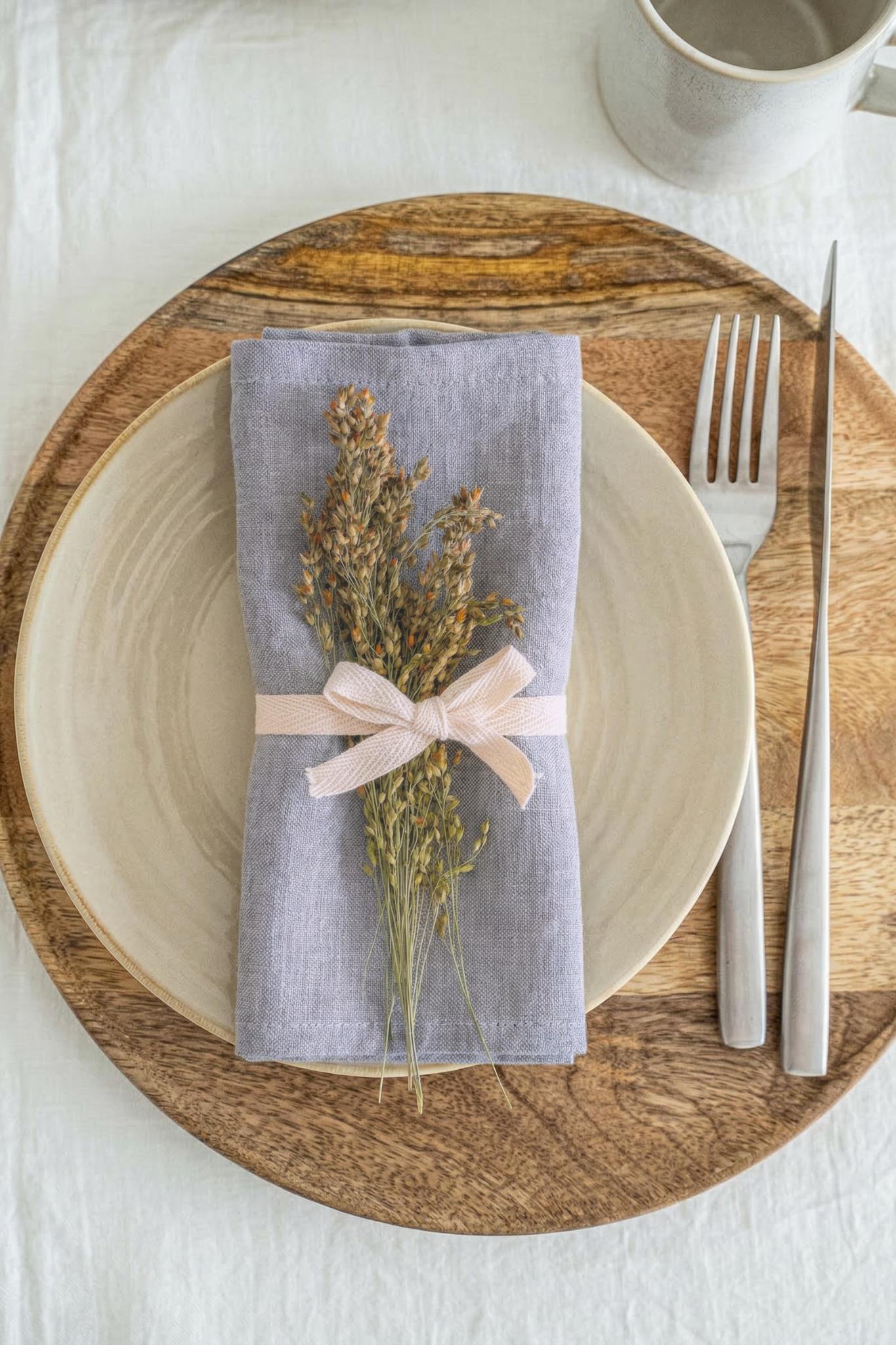 Dinner setting with a wooden plate, white bowl, gray napkin with dried flowers and a pink bow, and silver fork and knife on a white tablecloth.