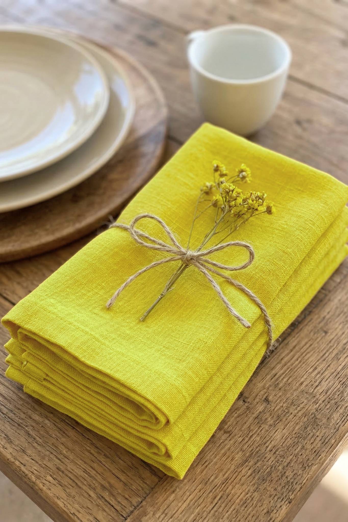 Yellow napkins tied with twine on a wooden table with plates and a cup in the background.
