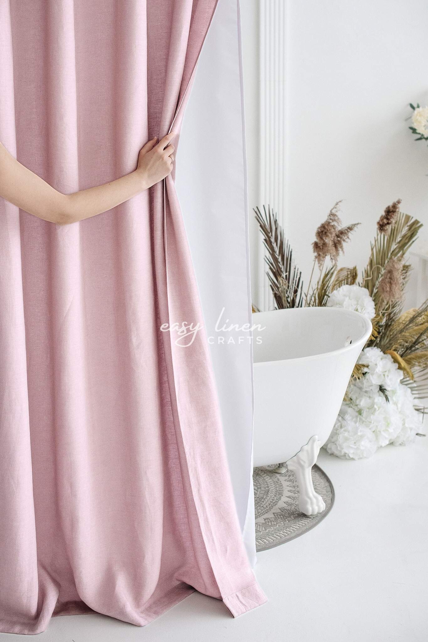 Person holding a pink linen shower curtain with a white bathtub and decorative plants in the background