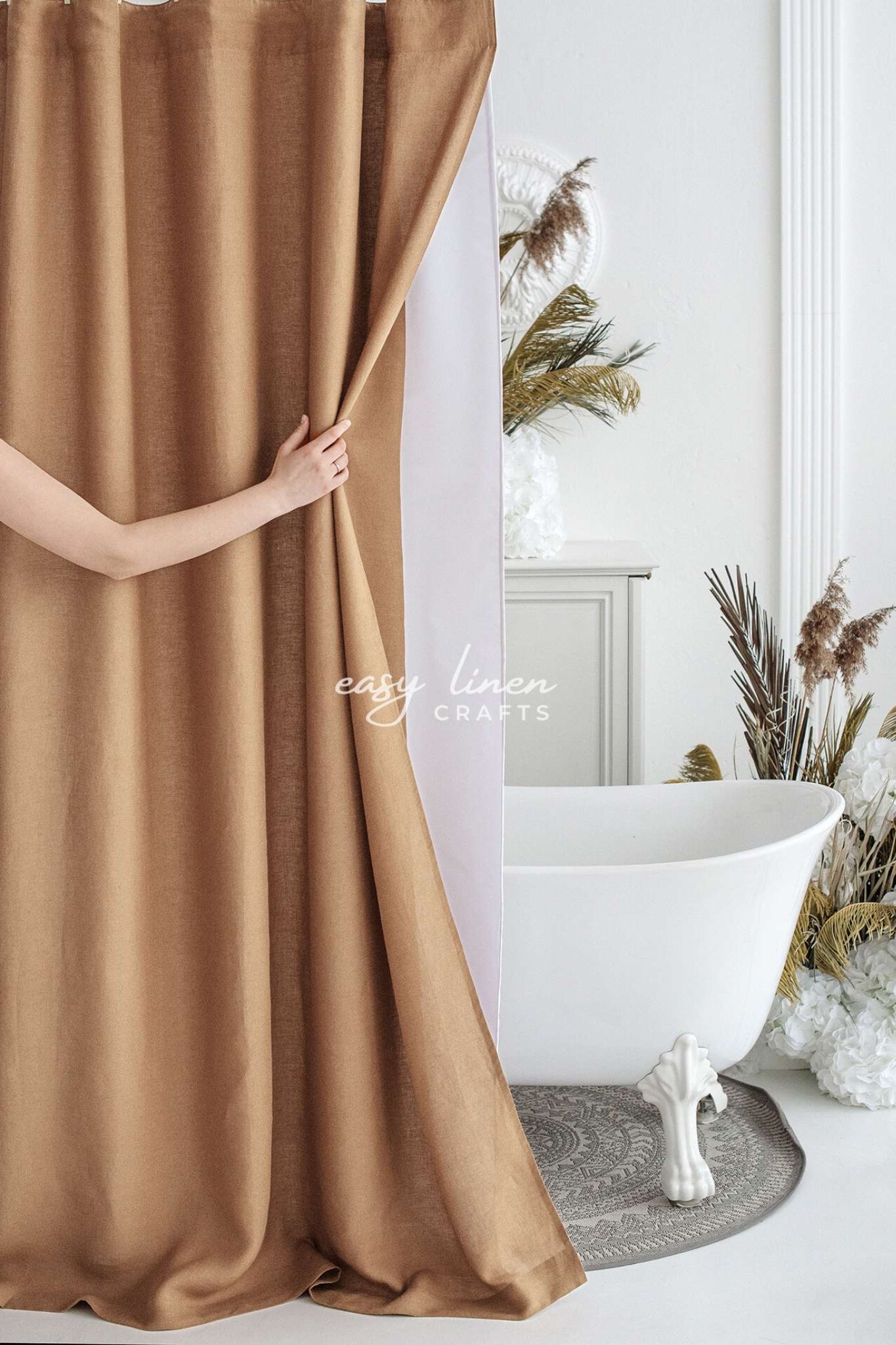 Camel Brown linen shower curtain being held open in a bathroom with a white bathtub and decorative plants.