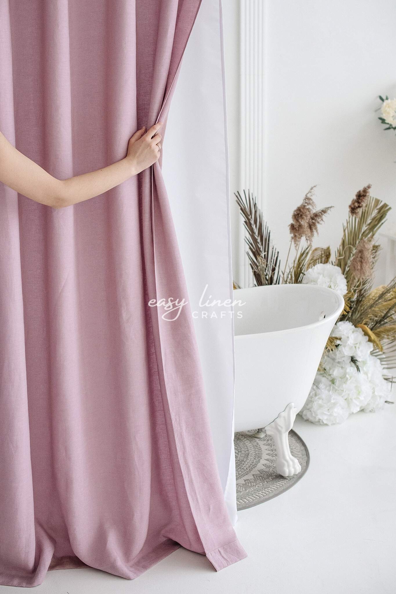 Person holding a dusty rose shower curtain with a bathtub and decorative plants in the background