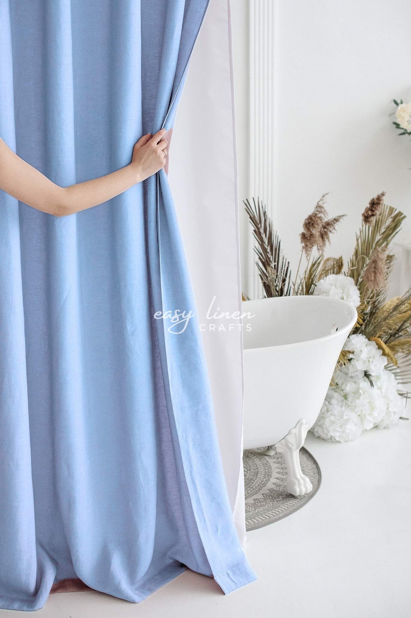 Person holding a light blue shower curtain in a bathroom with a bathtub and decorative plants.