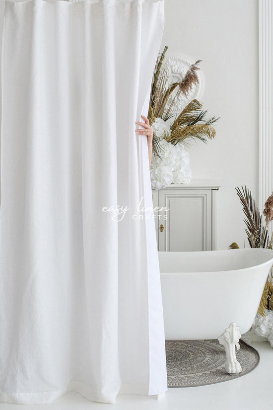 White linen shower curtain being pulled back to reveal a bathroom with a freestanding bathtub and decorative elements.