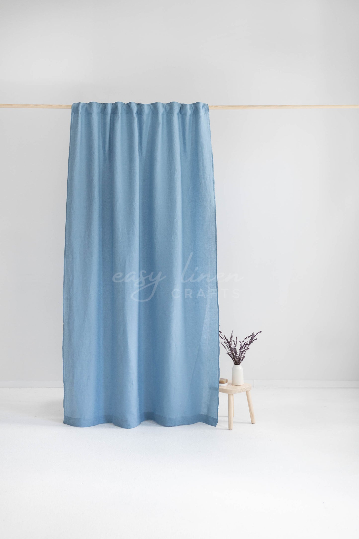 A light blue curtain panel hanging from a thin wooden rod against a clean white studio background, with a small wooden stool and vase of dried stems beside it. A faint 'easy linen curtains' watermark is visible across the center.
