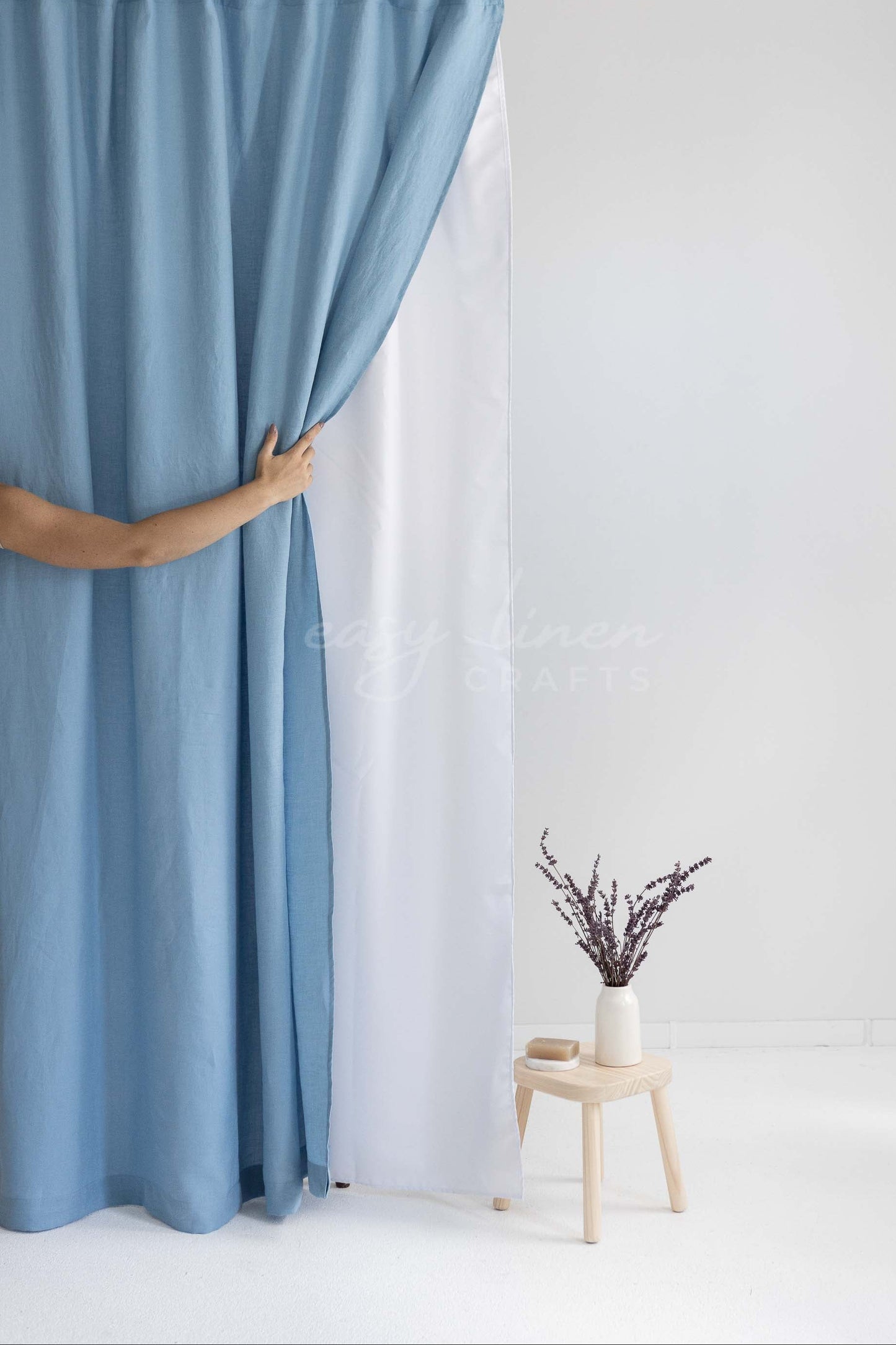 A person's hand pulls open a light blue curtain to reveal a white inner curtain panel hanging from a wooden rod. A small wooden stool with a vase of dried branches sits on the floor against a white wall.