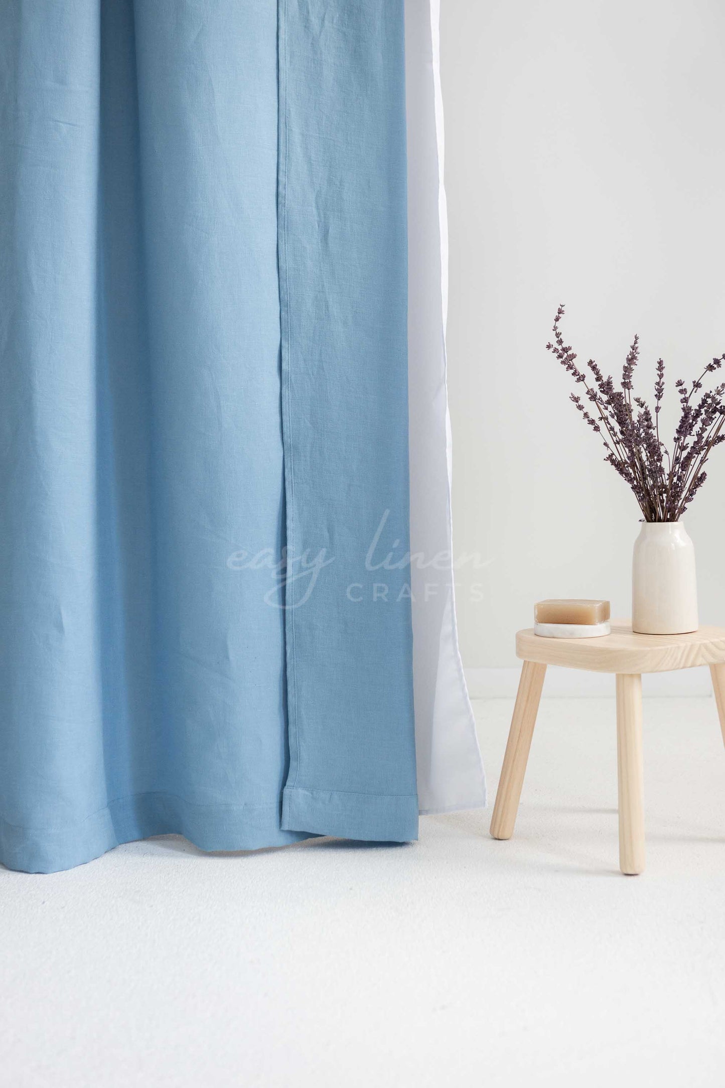 Light blue linen curtains hanging next to a white inner curtain, styled in a minimalist room with a small wooden stool holding a vase of dried flowers.