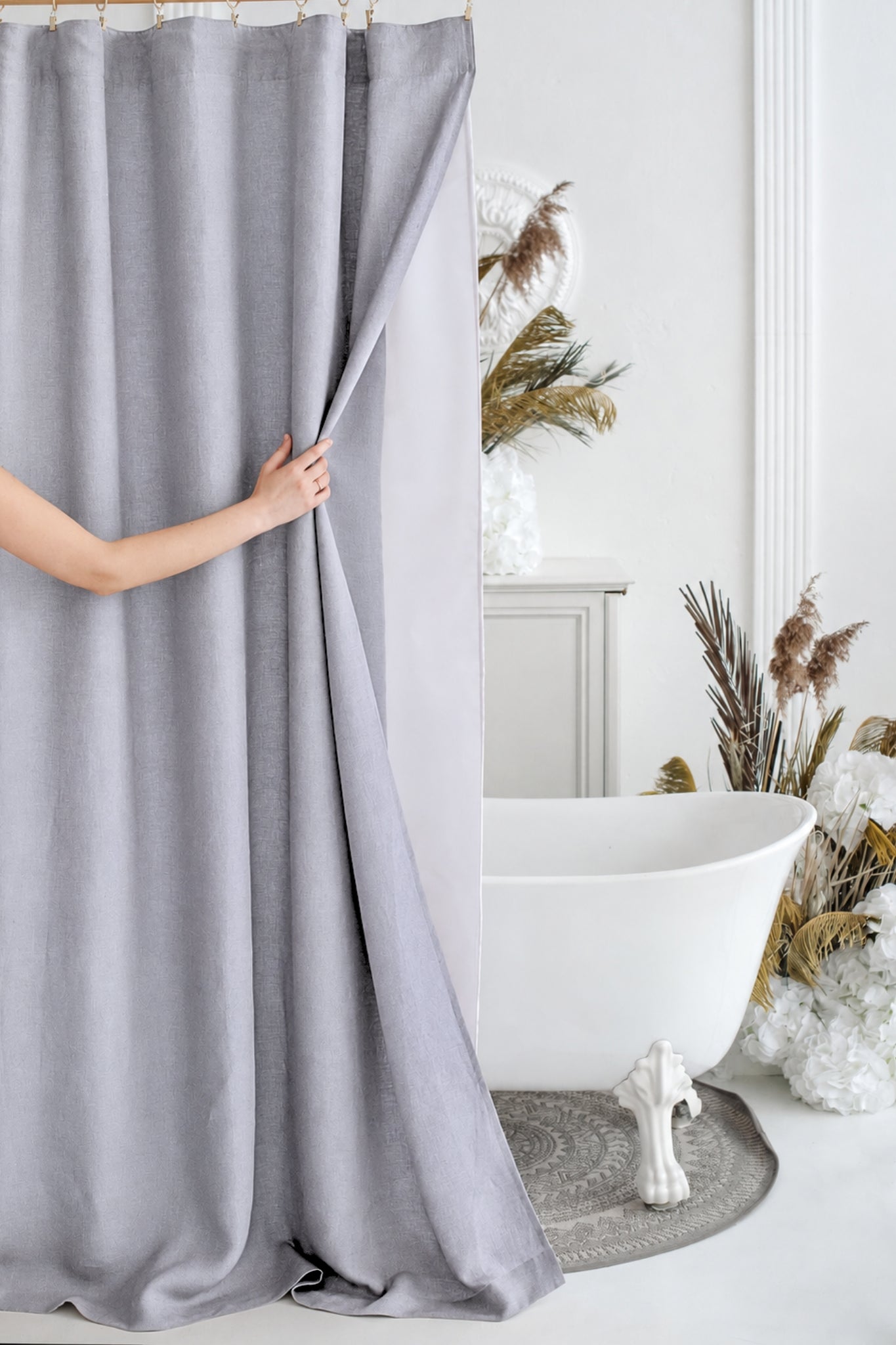 A person's arm and hand are shown pulling a light gray fabric shower curtain in a bright, modern bathroom setting next to a white clawfoot bathtub.