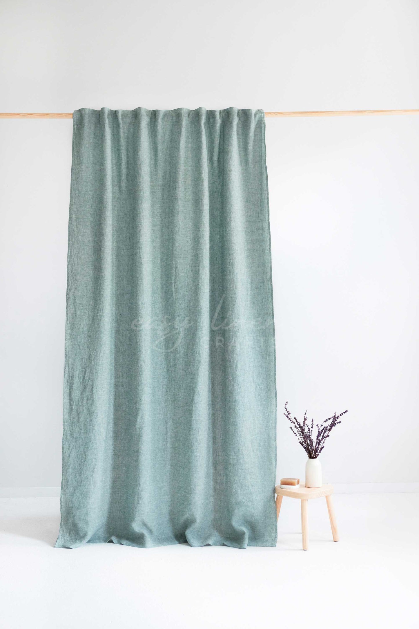 A sage green curtain panel hanging from a light wood rod against a white wall, with a small wooden stool and vase holding dried stems at the lower right.