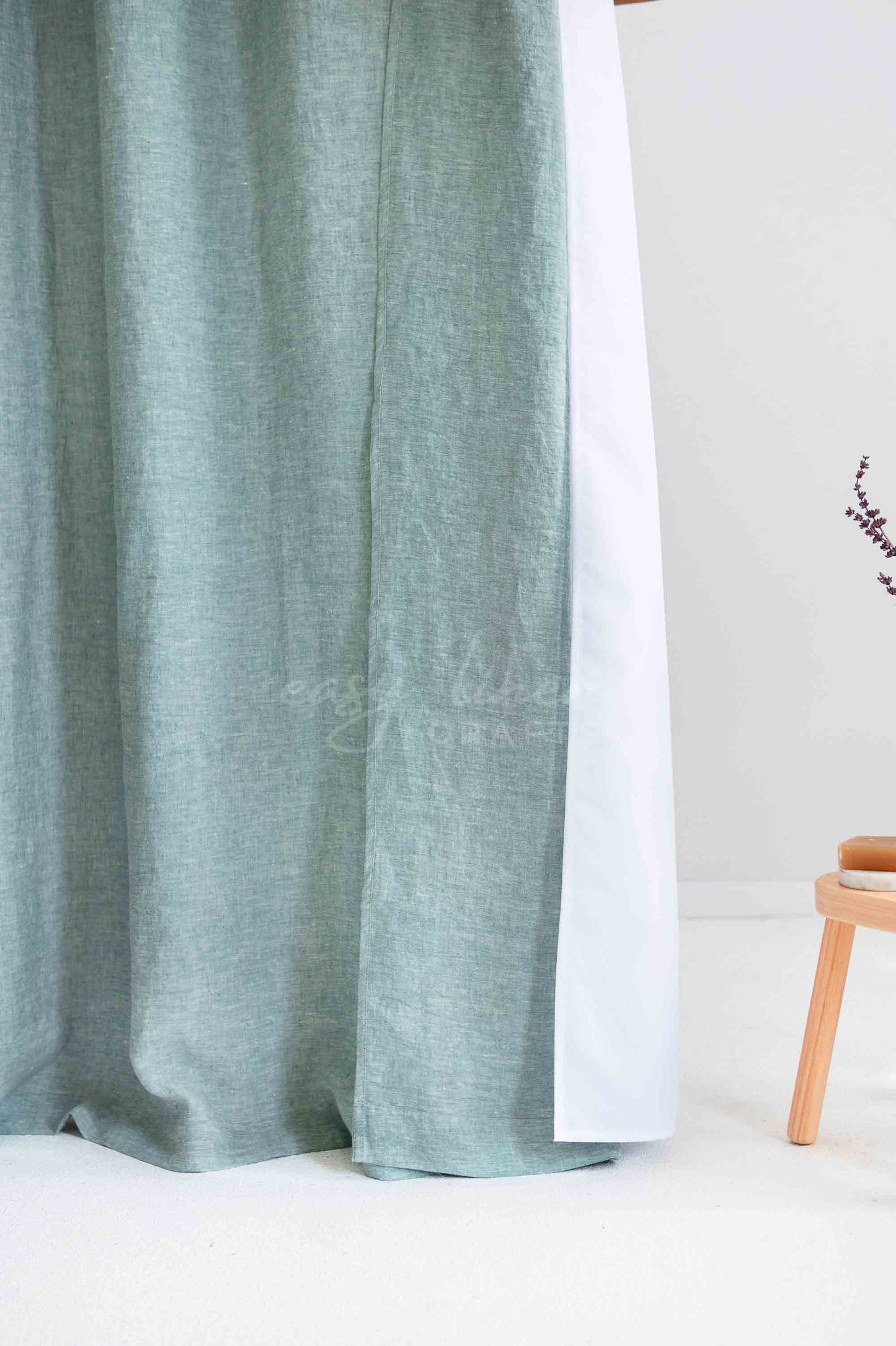A melange green linen curtain panel hanging next to a white backing or inner panel, with a small wooden stool partially visible on the right against a light wall.