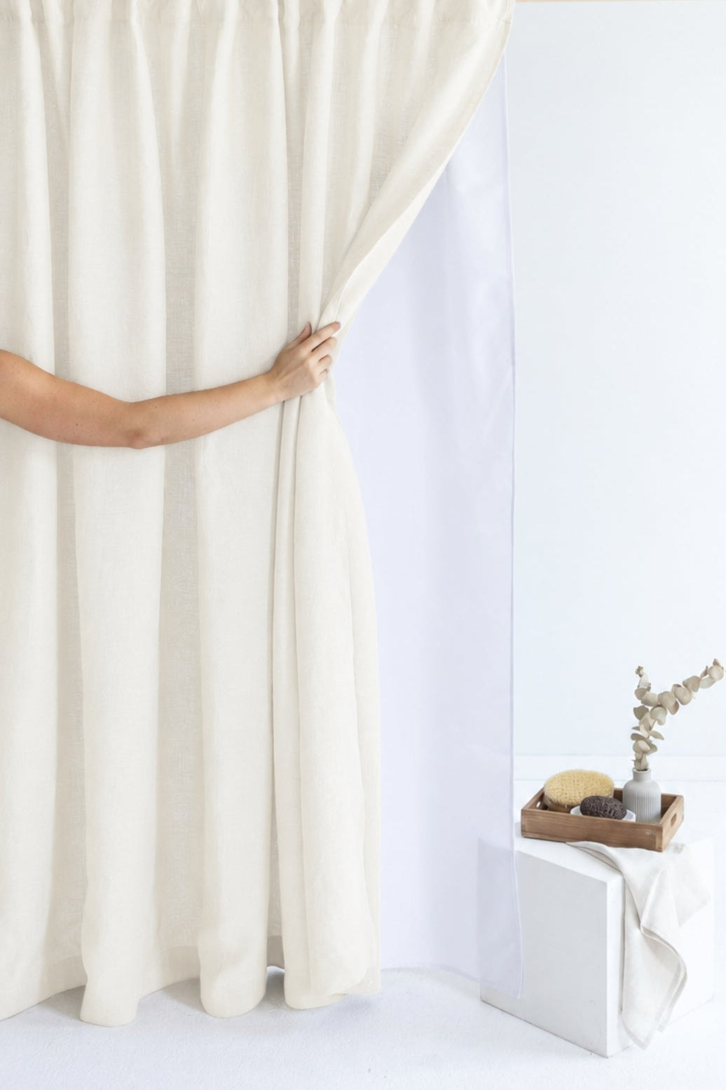 An off-white curtain panel being held open by a person's hand, revealing a white studio backdrop. A small white pedestal with a decorative tray and dried stems sits in the lower right corner against a clean white wall.