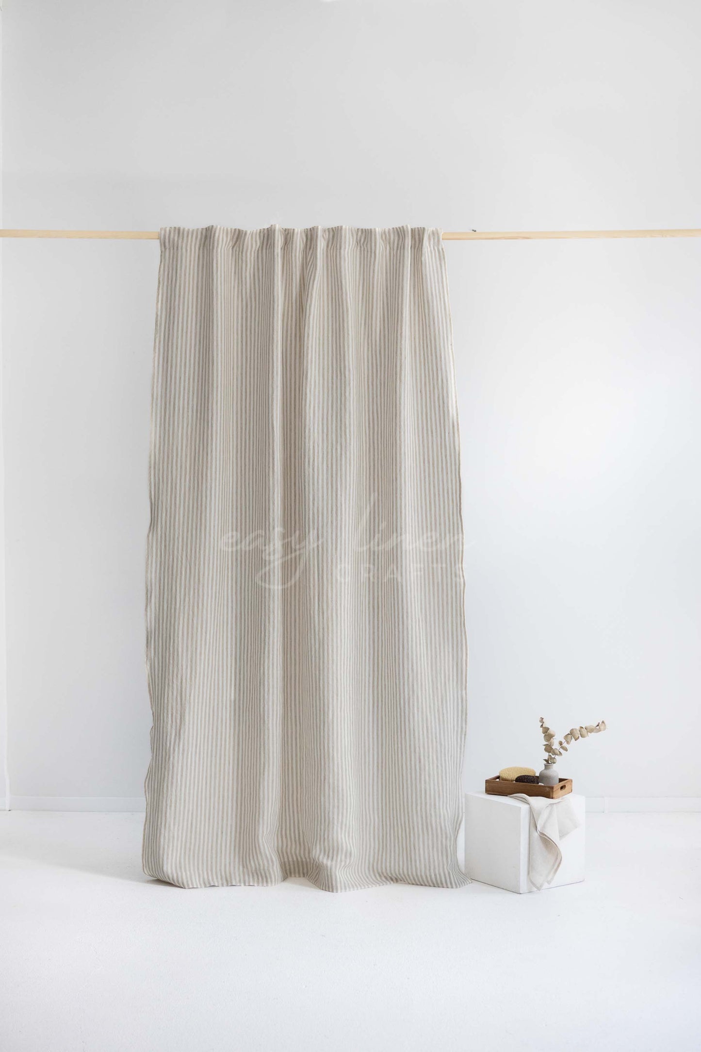 A light beige pleated curtain hanging from a thin wooden rod against a white wall, with a small decorative box and dried stems on the floor nearby.
