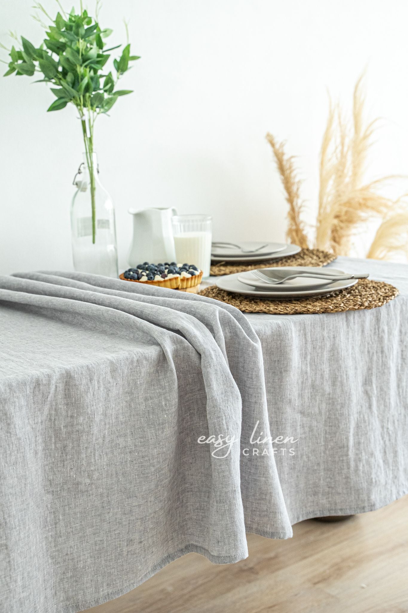 Table setting with a gray linen tablecloth, glass of milk, blueberries, and a vase with greenery.