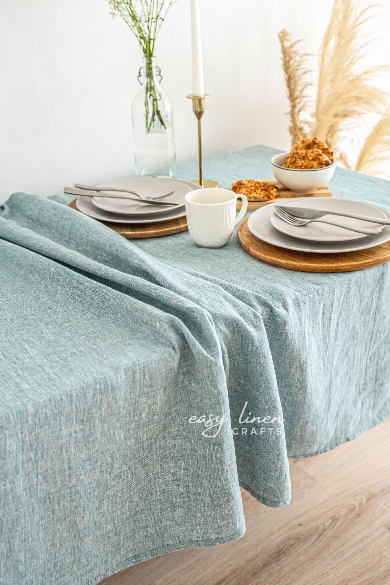 Table setting with melange green linen tablecloth, plates, cups, and cutlery on a wooden table.