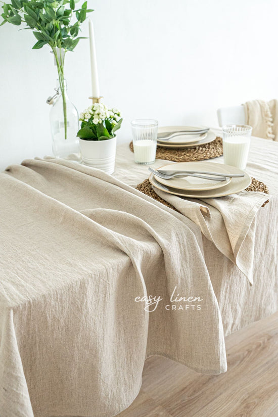 Beige linen tablecloth on a table with white plates, silverware, and greenery.