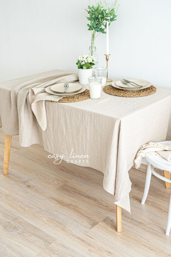 Beige linen tablecloth on a dining table with white chairs, glasses, and decorative items.