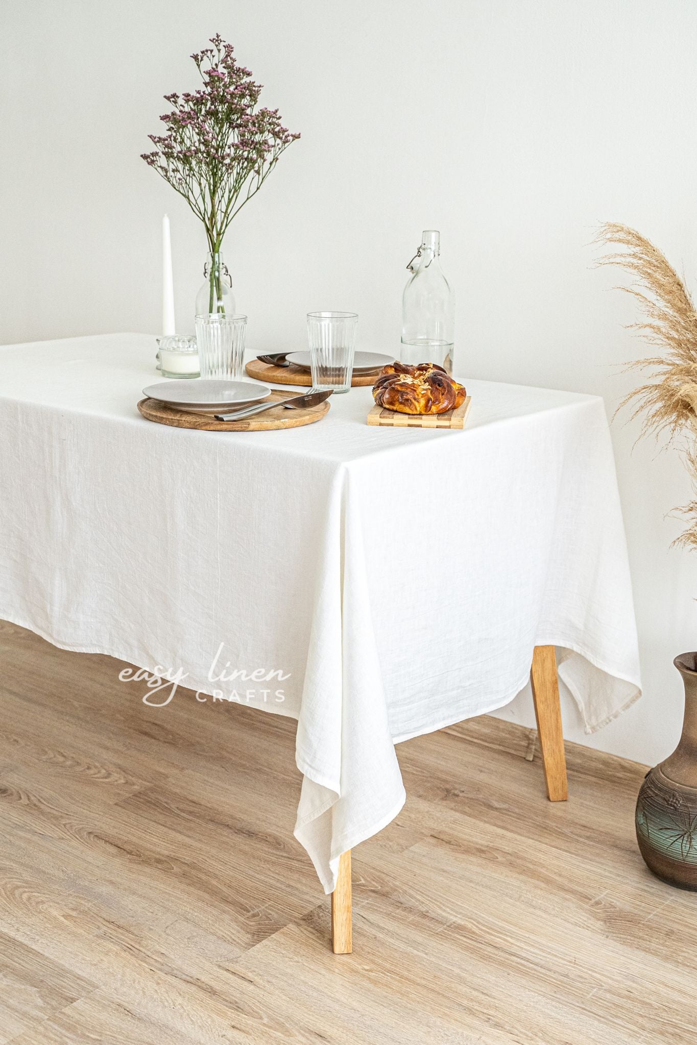Dining table set with an ivory linen tablecloth, plates, glasses, and a loaf of bread on a wooden floor.