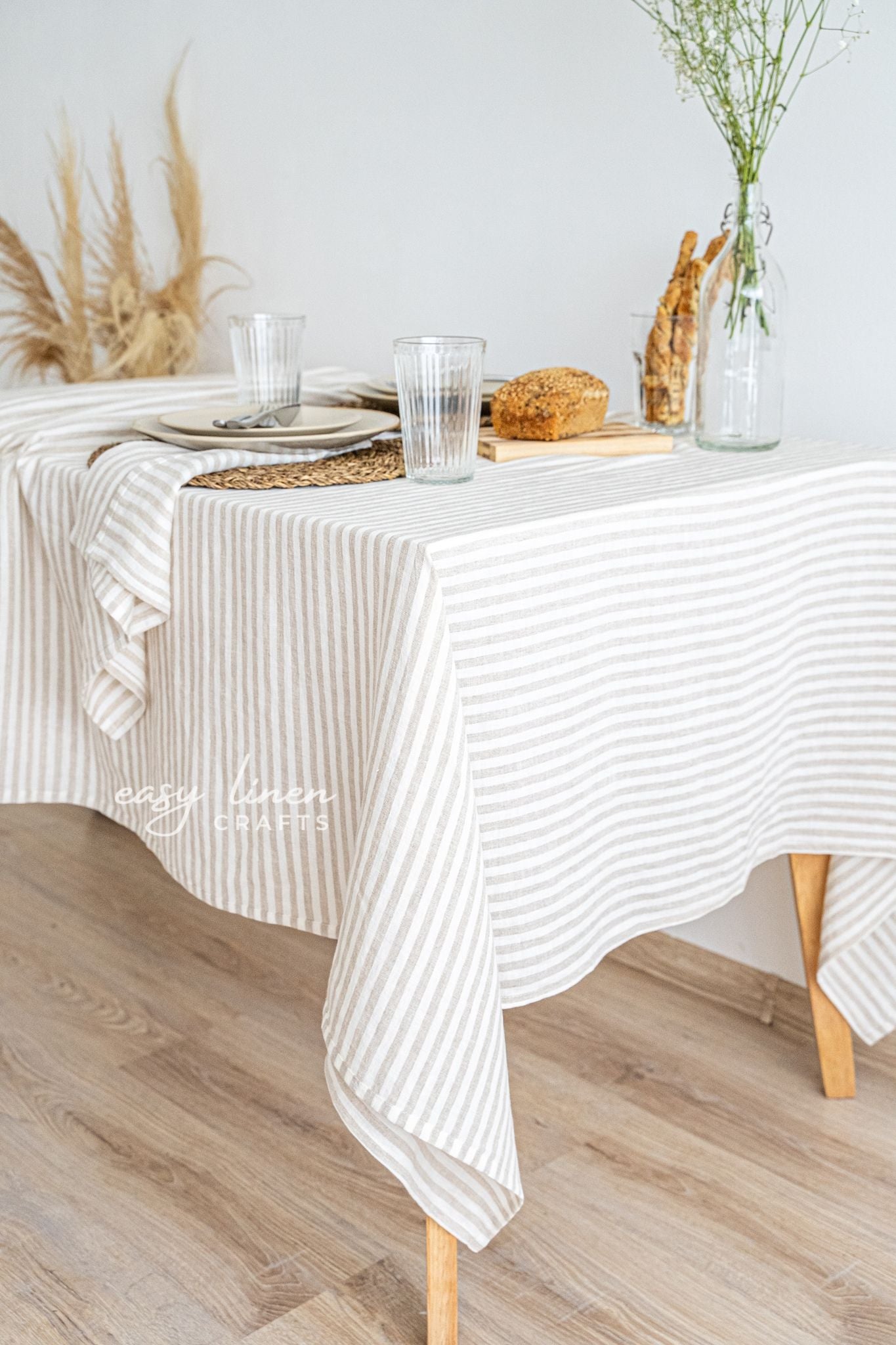 Dining table with a linen striped tablecloth, glasses, bread, and a vase on a wooden floor.