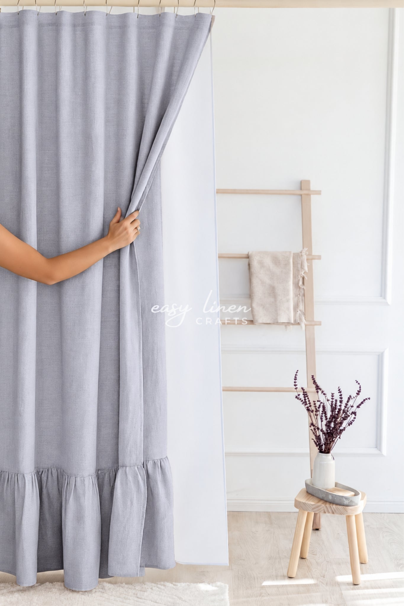 A light gray linen curtain panel with a ruffled hem, being held open by a person's hand in a bright, modern room. A wooden ladder shelf with a beige towel and a small stool with dried lavender are visible in the background.