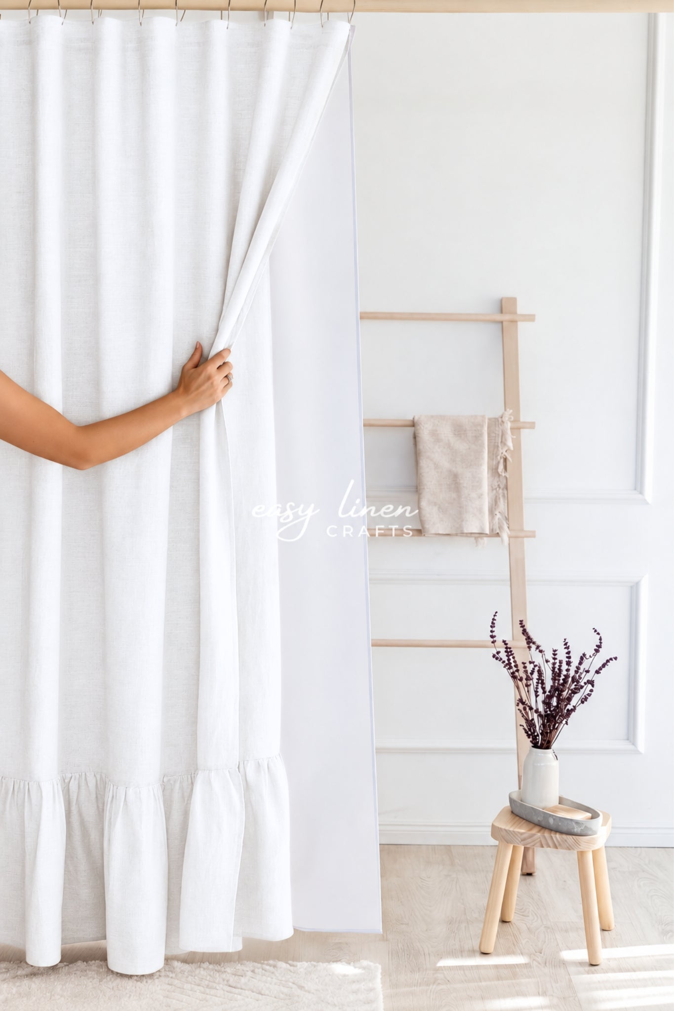 A white curtain panel with a ruffled hem is being held open by a person's hand in a bright, modern room. A wooden ladder shelf with a beige towel and a small stool with dried flowers are visible in the background.