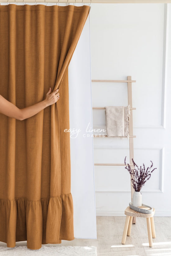 Person opening a mustard yellow shower curtain in a room with a wooden stool and plant.