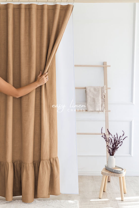 Brown shower curtain being pulled open in a room with a wooden stool and plant.