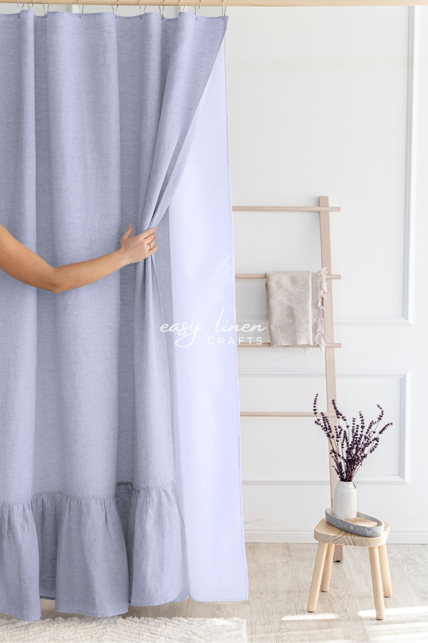 Person holding a light gray shower curtain in a bathroom with a wooden stool and plant.