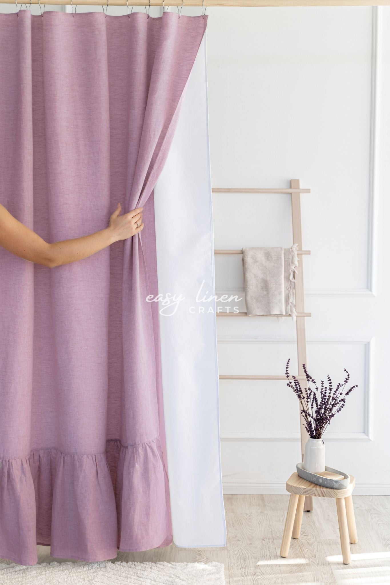 Person holding a dusty rose shower curtain in a room with a wooden stool and decorative items.