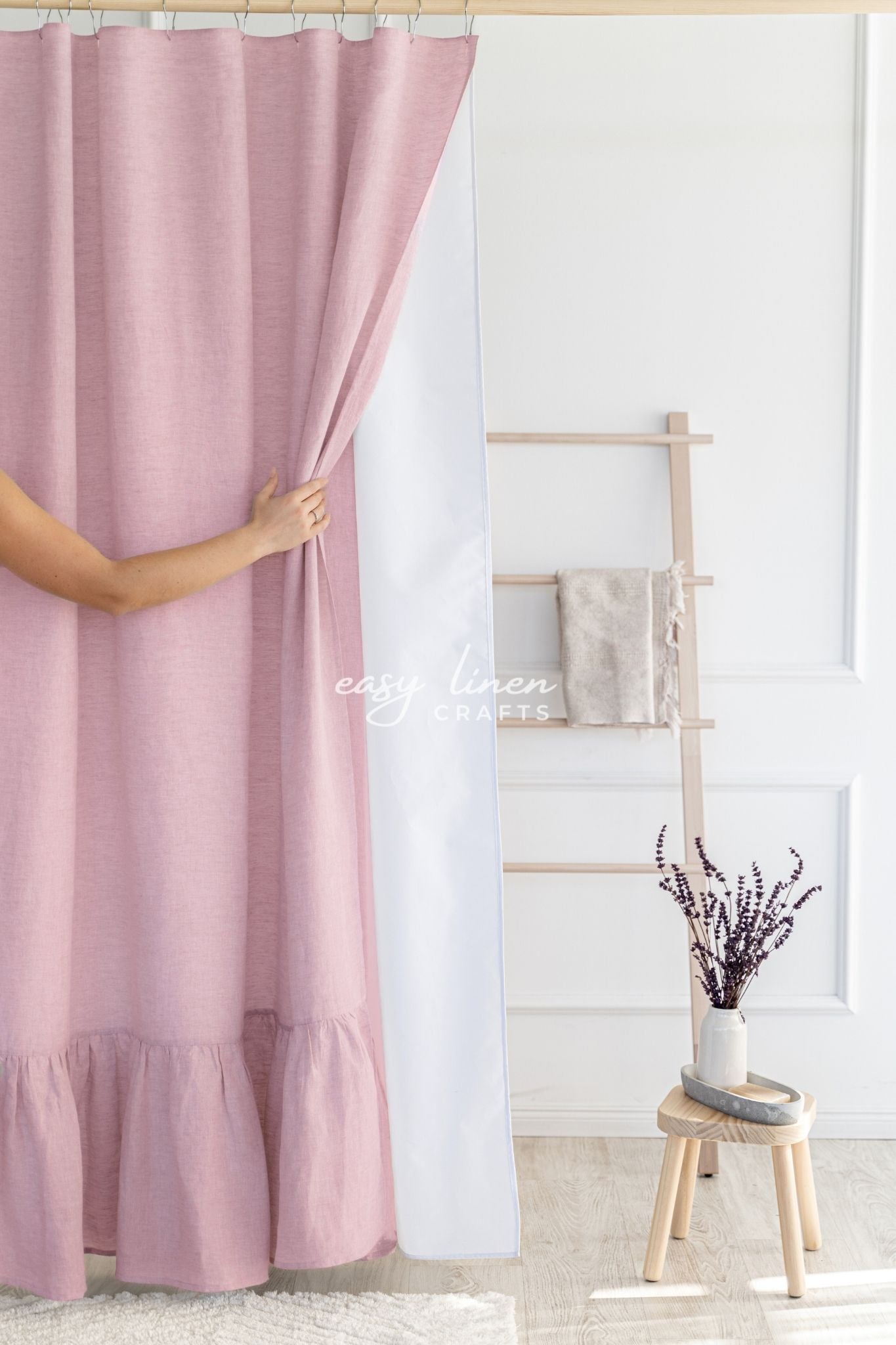 Pink linen shower curtain being adjusted in a room with a wooden stool and decorative items.