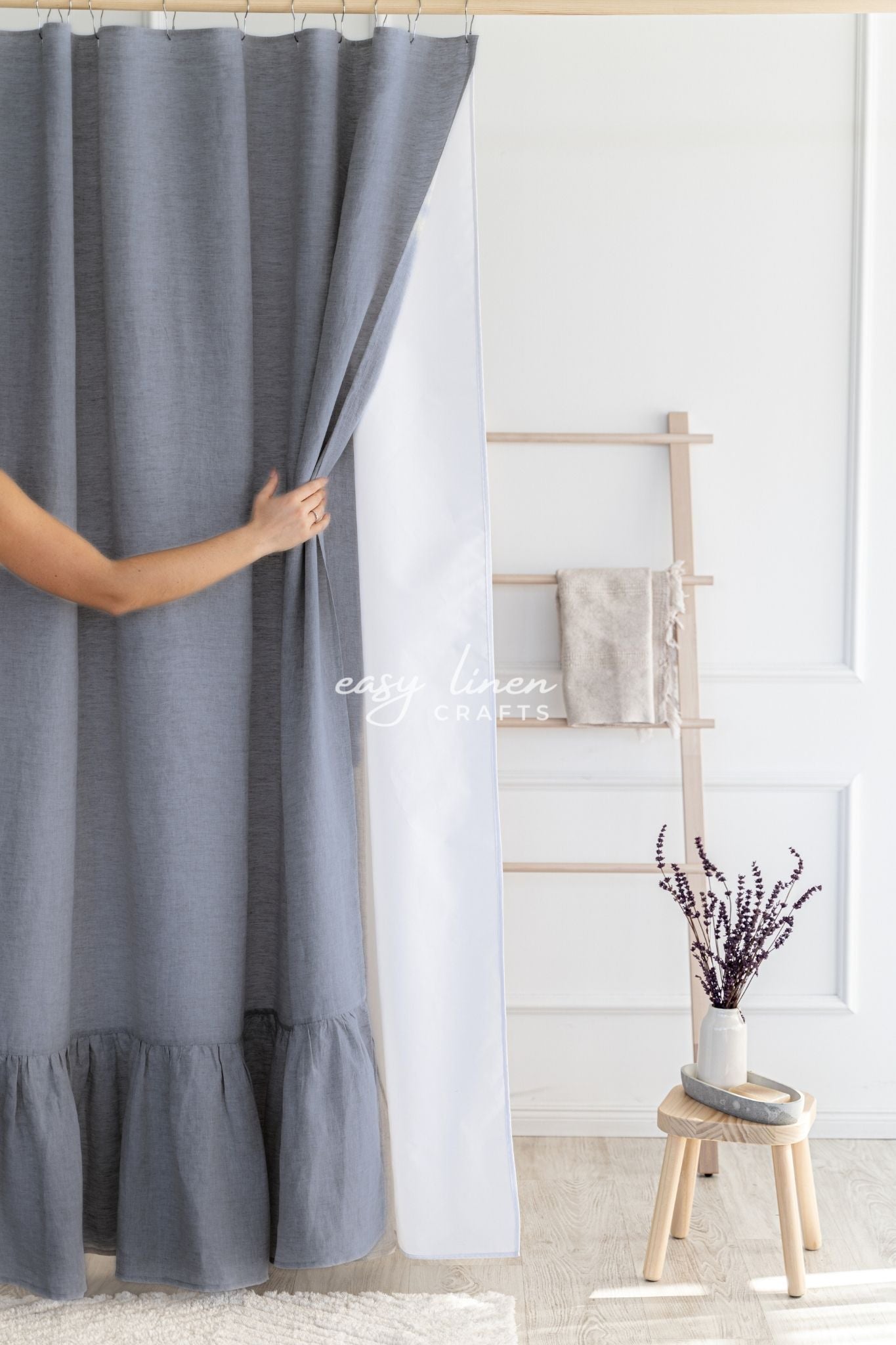 Person adjusting a graphite gray linen shower curtain in a room with a wooden stool and decorative items.