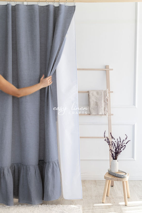Person adjusting a graphite gray linen shower curtain in a room with a wooden stool and decorative items.